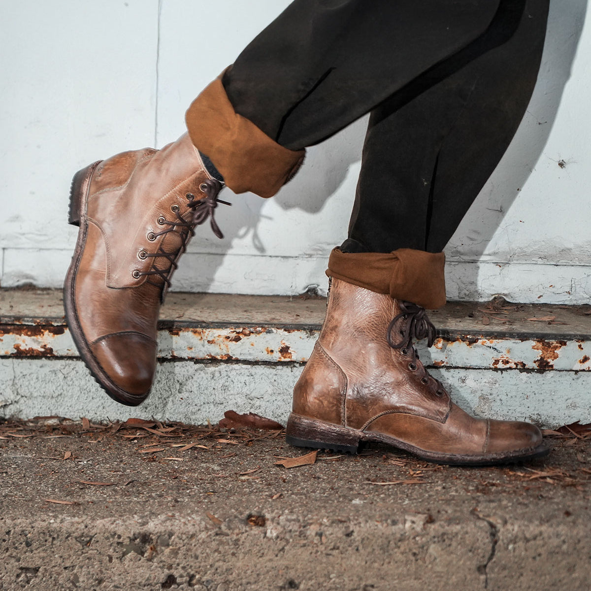 A person wearing the Bed|Stü Protege brown leather lace-up boots with a classic Goodyear welt and rolled-up black pants, standing on a concrete surface against a weathered metal background.