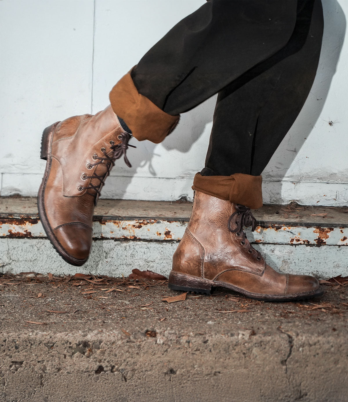 A person wearing the Bed|Stü Protege brown leather lace-up boots with a classic Goodyear welt and rolled-up black pants, standing on a concrete surface against a weathered metal background.