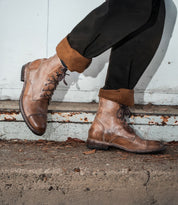 A person wearing the Bed|Stü Protege brown leather lace-up boots with a classic Goodyear welt and rolled-up black pants, standing on a concrete surface against a weathered metal background.
