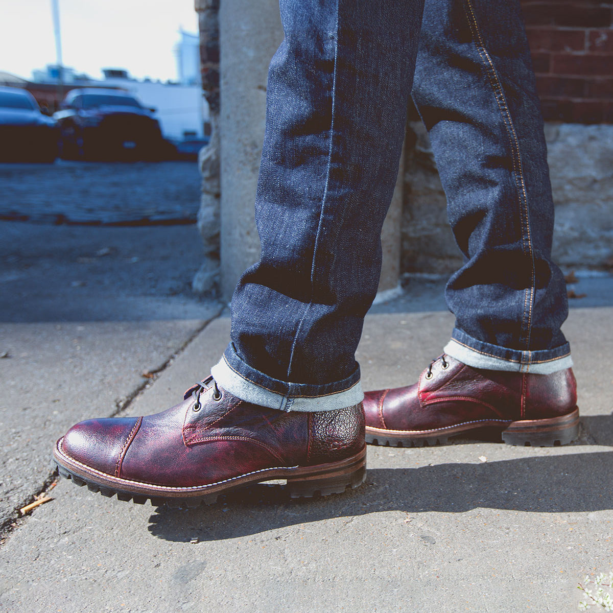 A person wearing dark blue jeans and burgundy leather Bed|Stü Protege Trek Goodyear welt shoes stands on a sidewalk next to a brick wall on a sunny day.