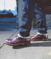 A person wearing dark blue jeans and burgundy leather Bed|Stü Protege Trek Goodyear welt shoes stands on a sidewalk next to a brick wall on a sunny day.