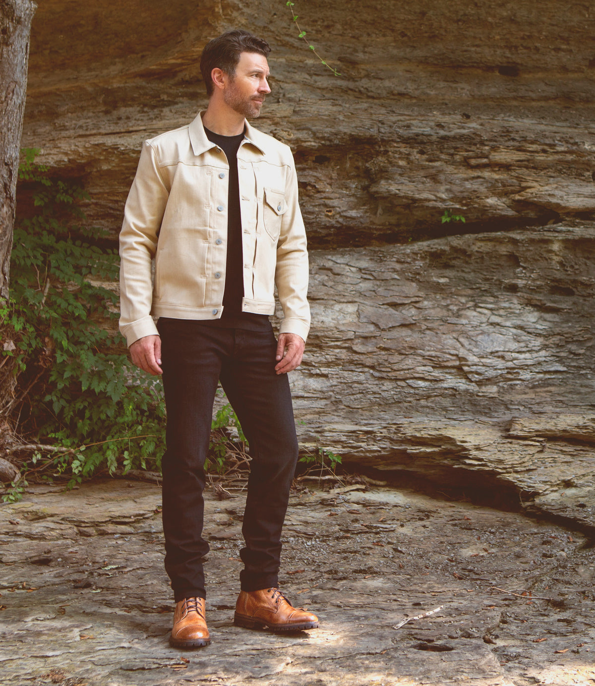 A man stands outdoors on rocky ground, wearing the Bed|Stü Protege Trek boots, paired with a cream-colored jacket and black pants, with a rocky wall and greenery in the background.