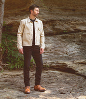 A man stands outdoors on rocky ground, wearing the Bed|Stü Protege Trek boots, paired with a cream-colored jacket and black pants, with a rocky wall and greenery in the background.