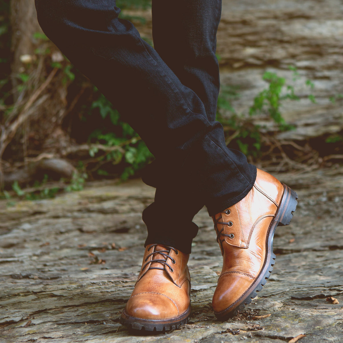 A person wearing dark pants and Bed|Stü Protege Trek tan leather lace-up combat boots—crafted with a Goodyear welt for extra durability—stands on a stone path with legs crossed, green plants visible in the background.