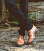 A person wearing dark pants and Bed|Stü Protege Trek tan leather lace-up combat boots—crafted with a Goodyear welt for extra durability—stands on a stone path with legs crossed, green plants visible in the background.