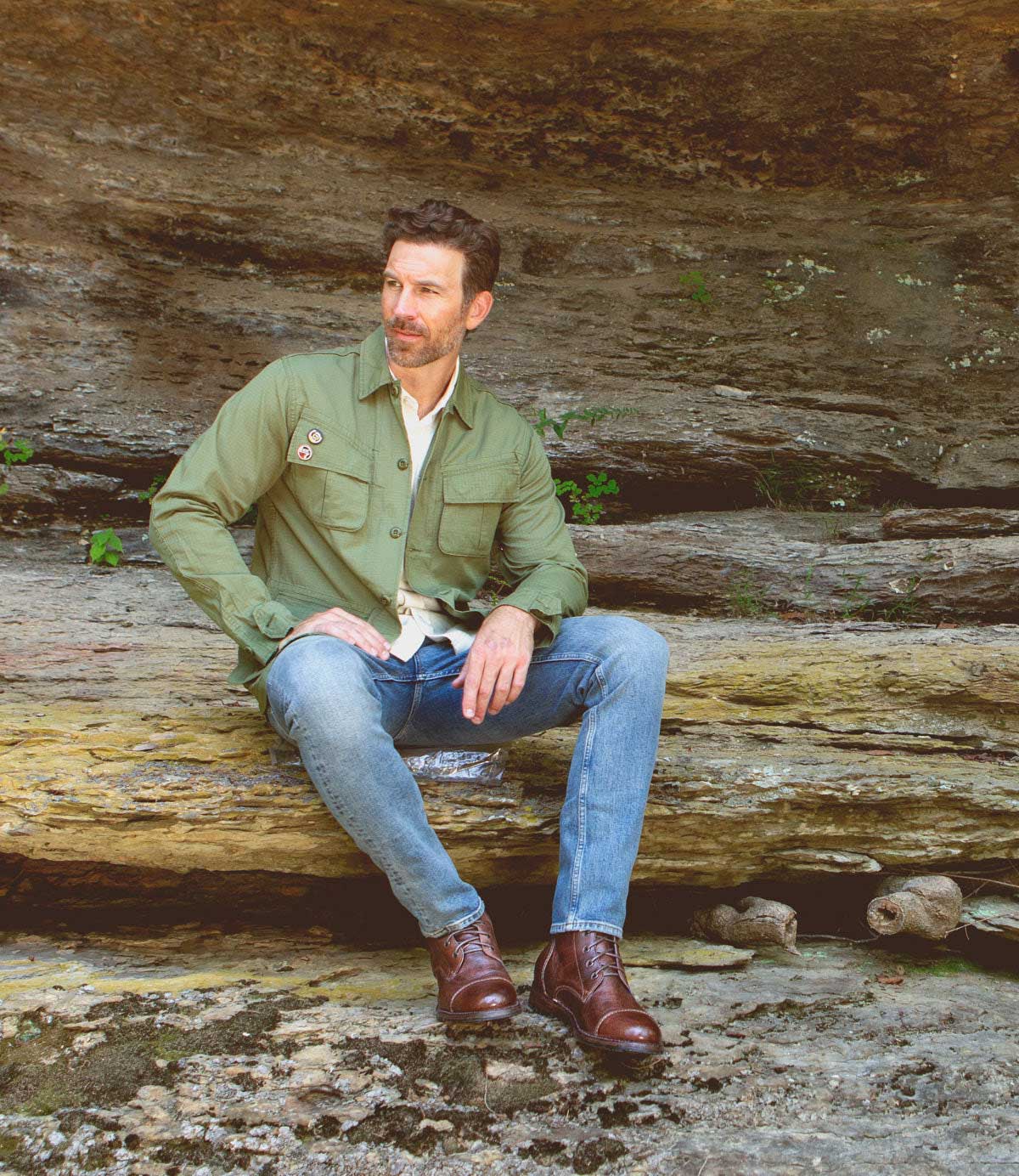 A man wearing the Bed|Stü Protege boots, along with a green jacket and jeans, sits on a rock ledge outdoors with a stone wall behind him.