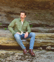 A man wearing the Bed|Stü Protege boots, along with a green jacket and jeans, sits on a rock ledge outdoors with a stone wall behind him.