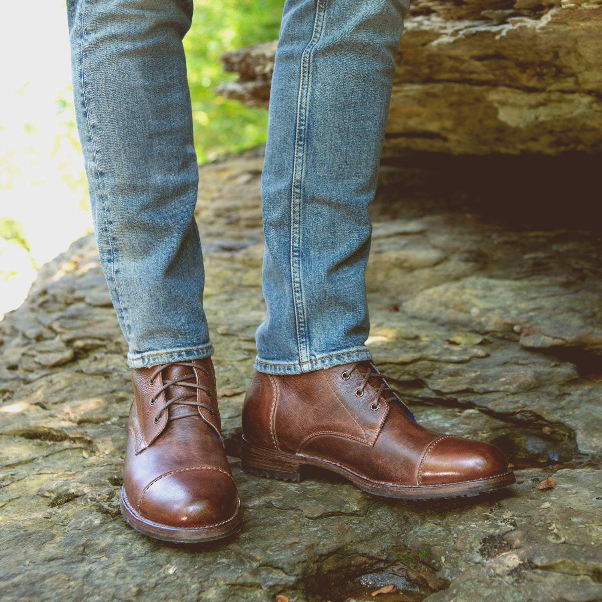 A person wearing blue jeans and durable Bed|Stü Protege boots stands on a rocky surface outdoors, with greenery visible in the background. The brown leather Protege boots showcase classic Goodyear welt construction, making them perfect for rugged adventures.