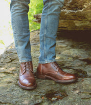 A person wearing blue jeans and durable Bed|Stü Protege boots stands on a rocky surface outdoors, with greenery visible in the background. The brown leather Protege boots showcase classic Goodyear welt construction, making them perfect for rugged adventures.
