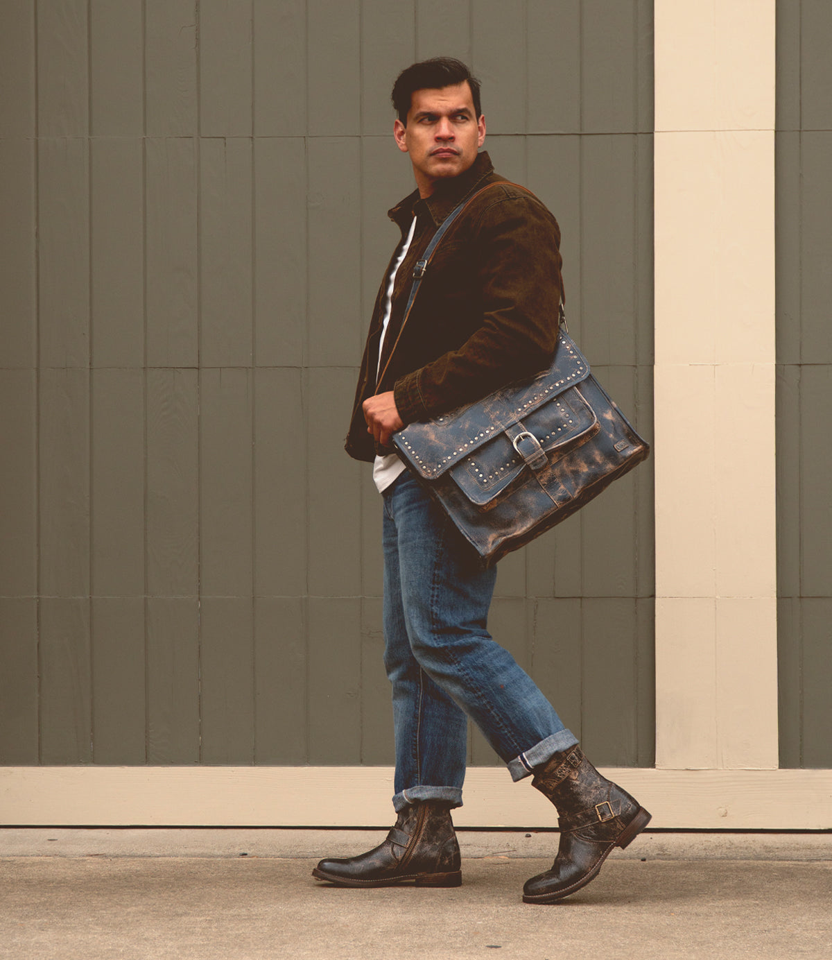 A man dressed in a brown jacket, blue jeans, and boots stands in front of a gray fence, carrying the Bed|Stü Pub travel bag that perfectly complements his style.