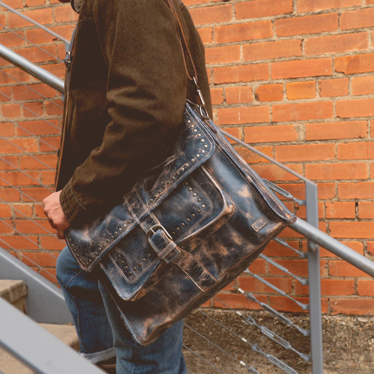 A person in a brown jacket carries the Bed|Stü Pub, a large, worn leather work bag, while walking up metal stairs next to a brick wall.