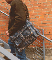 A person in a brown jacket carries the Bed|Stü Pub, a large, worn leather work bag, while walking up metal stairs next to a brick wall.
