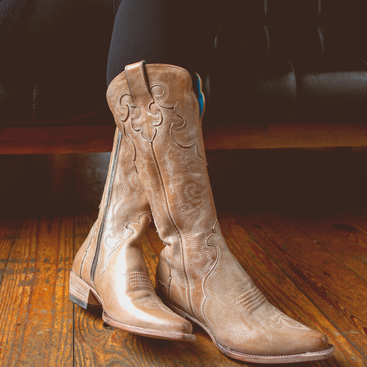 A person wearing the "Queen" Western boots by Bed|Stü, crafted from vegetable-tanned leather in a tan hue, sits on a wooden floor with legs crossed; the boots showcase pointed toes, stacked heels, and delicate Fleur-de-lis embroidery.