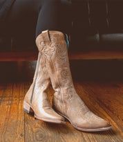 A person wearing the "Queen" Western boots by Bed|Stü, crafted from vegetable-tanned leather in a tan hue, sits on a wooden floor with legs crossed; the boots showcase pointed toes, stacked heels, and delicate Fleur-de-lis embroidery.