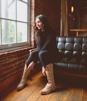 A woman in a black outfit and the Queen tan Western boots by Bed|Stü, featuring subtle Fleur-de-lis details, sits on a black sofa by a window in a room with brick walls and wooden floors.