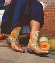 A person in a blue denim skirt sits on a white chair, wearing Bed|Stü’s Raise short boots in tan and orange with snip toe detailing. Cantaloupes are on the floor beside them against a brick wall.
