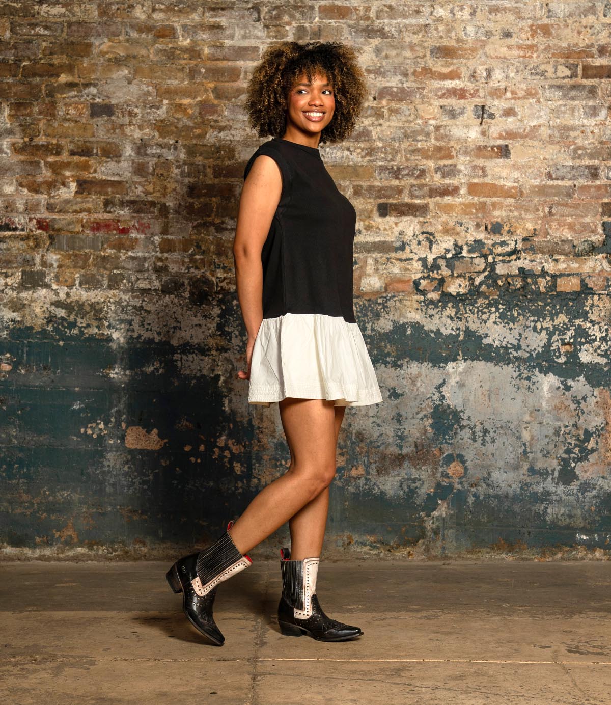 A woman with curly hair, dressed in a black sleeveless top and a white skirt, stands against a worn brick wall smiling at the camera, wearing Bed|Stü's Raise black Western short boots with a snip toe.