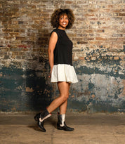 A woman with curly hair, dressed in a black sleeveless top and a white skirt, stands against a worn brick wall smiling at the camera, wearing Bed|Stü's Raise black Western short boots with a snip toe.