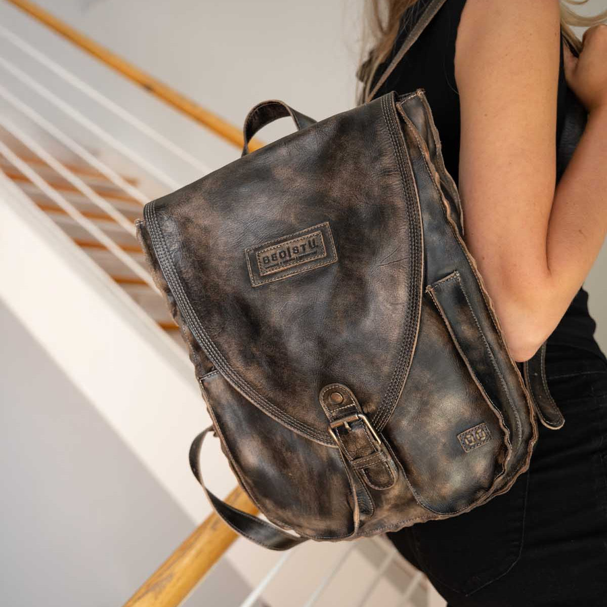 A person in black clothing stands on stairs, carrying the Ranger backpack by Bed|Stü—a sturdy, worn brown leather bag with a visible logo on the flap—making it an ideal choice for everyday wear.
