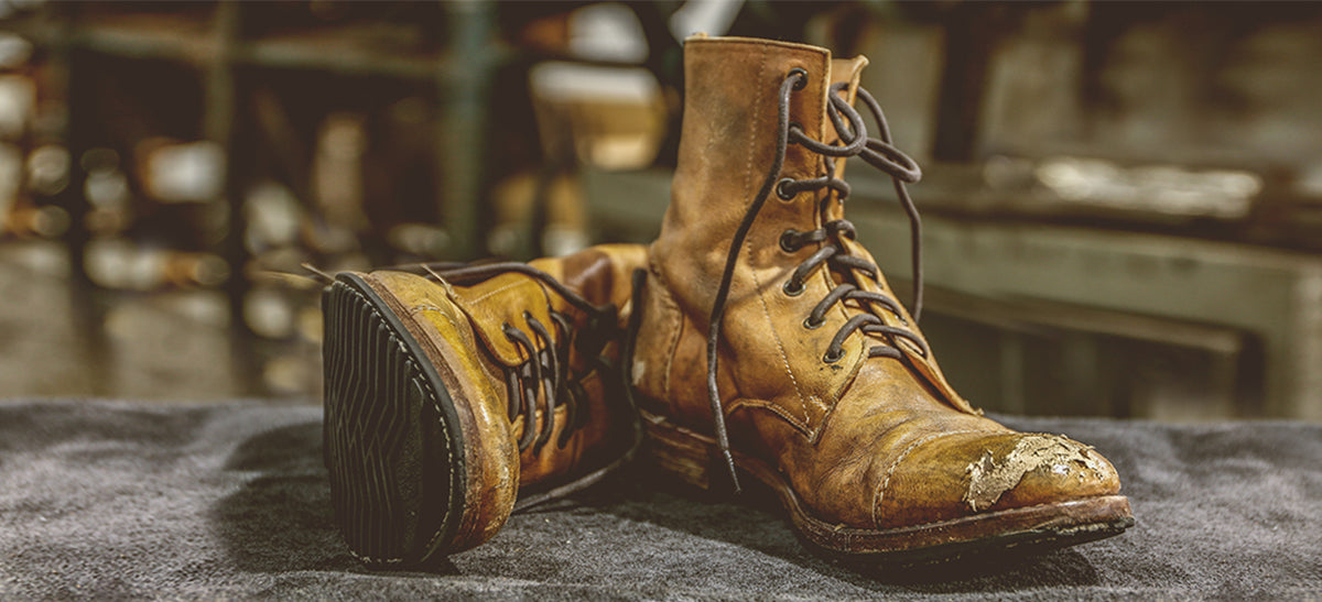 Worn tan leather lace-up boots on a dark cloth, one showing a patched and scuffed toe.