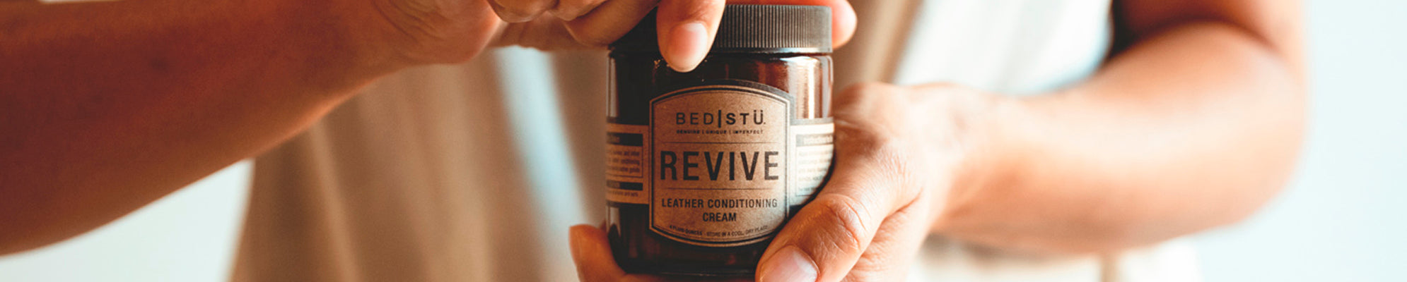 Hands holding a jar of BED|STU Revive leather conditioning cream.