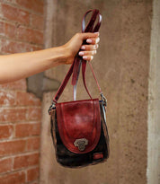 A hand holds up the Bed|Stü Rhyme, a compact crossbody bag crafted in two-tone brown and red leather with a metal clasp, against a brick and concrete background—offering a stylish hands-free accessory for any occasion.