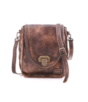 The Bed|Stü Rhyme is a brown leather crossbody bag with a vintage finish, featuring a metal clasp closure, adjustable strap for hands-free convenience, and decorative stitching, shown on a white background.