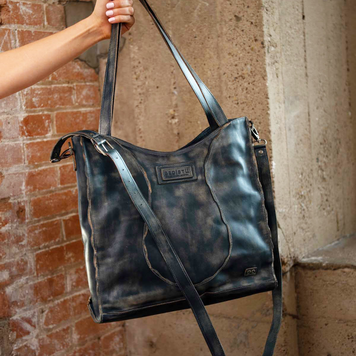 A hand holds the Bed|Stü Ridgefield tote, crafted from large, distressed black full-grain leather with two sturdy straps and a subtle embossed logo, set against brick and concrete walls. The stylish Ridgefield features a thoughtfully organized interior to keep daily essentials neatly in place.