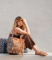 A woman with long blonde hair sits on a ledge against a light textured wall, wearing a brown outfit and sandals, holding the Bed|Stü Rimes handcrafted leather tote with fringe detail, with patterned cushions beside her.