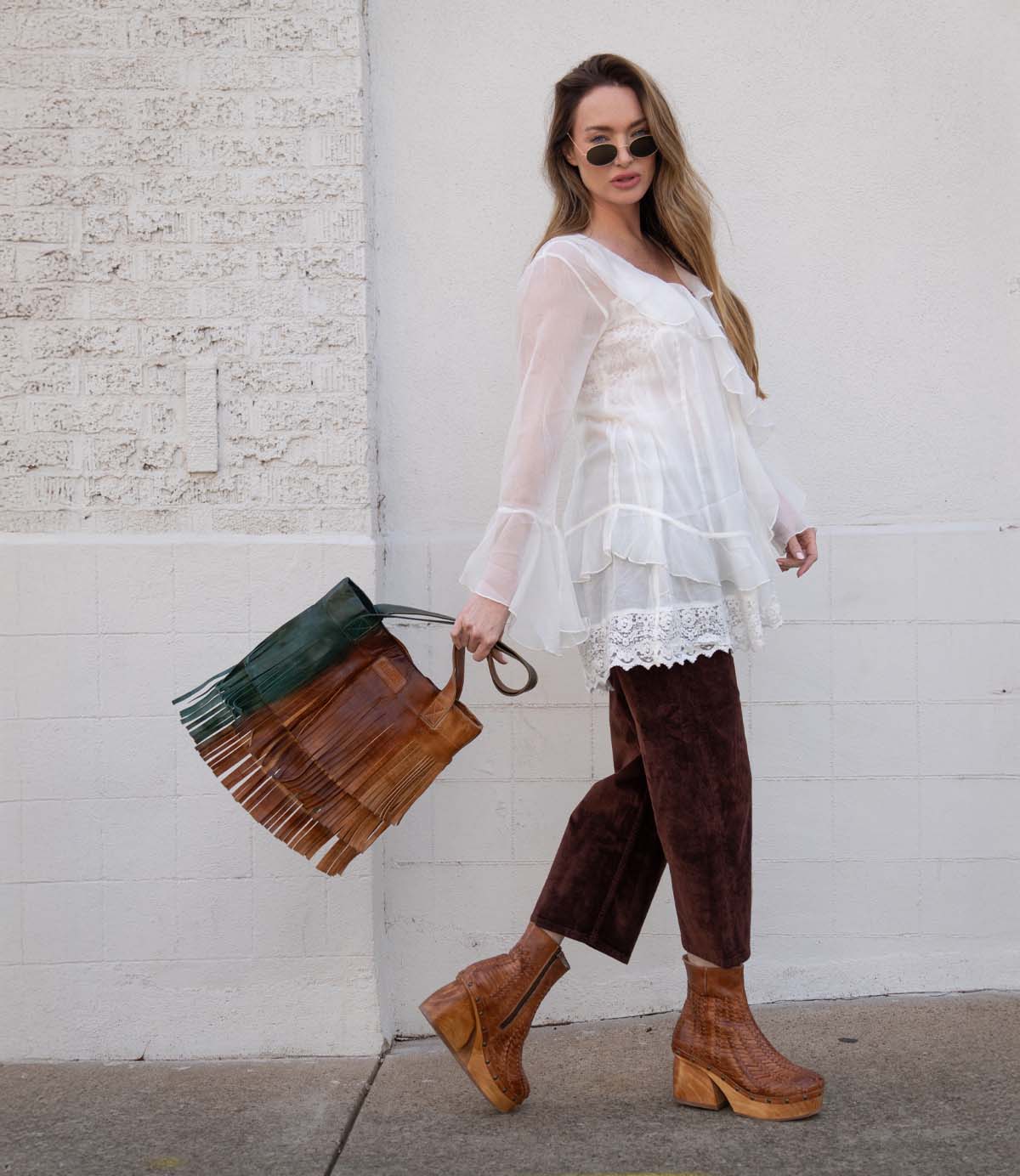 A woman wearing sunglasses, a sheer white blouse, brown suede pants, and brown platform boots stands against a white textured wall, holding the Bed|Stü Rimes handcrafted leather tote featuring fringe detail and a roomy interior.