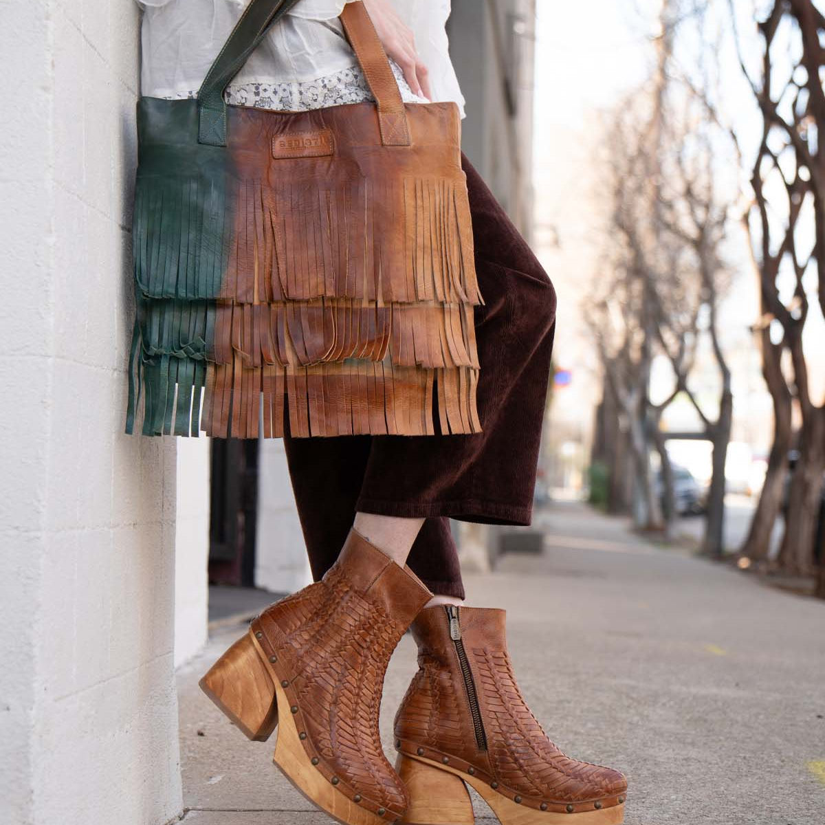 A person wearing the Rimes brown woven platform boots by Bed|Stü holds a two-tone leather fringed handbag with striking fringe detail while standing on a sidewalk next to a white wall.