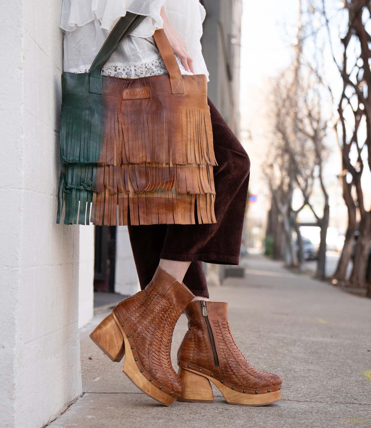 A person wearing the Rimes brown woven platform boots by Bed|Stü holds a two-tone leather fringed handbag with striking fringe detail while standing on a sidewalk next to a white wall.