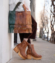 A person wearing the Rimes brown woven platform boots by Bed|Stü holds a two-tone leather fringed handbag with striking fringe detail while standing on a sidewalk next to a white wall.