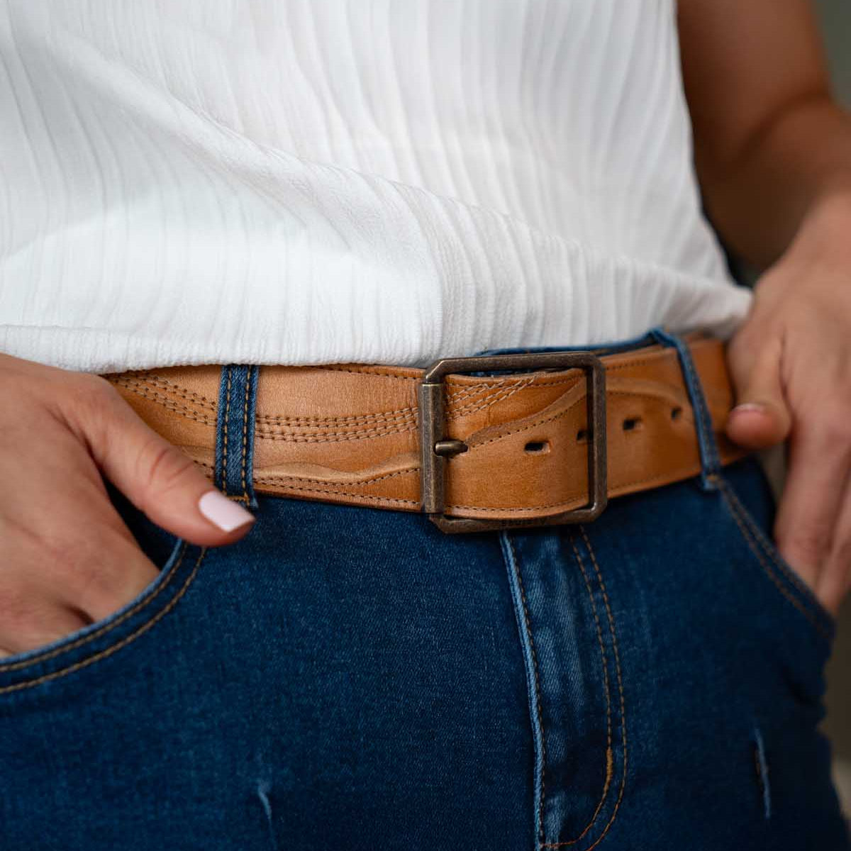A person wearing a white textured top, blue jeans, and the Bed|Stü Riven heritage leather belt with a rectangular buckle, with hands in pockets.