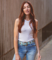 A woman with long red hair stands outdoors next to a textured concrete wall, wearing the Bed|Stü Roam patterned belt with geometric designs, styled with a white sleeveless top and blue jeans.