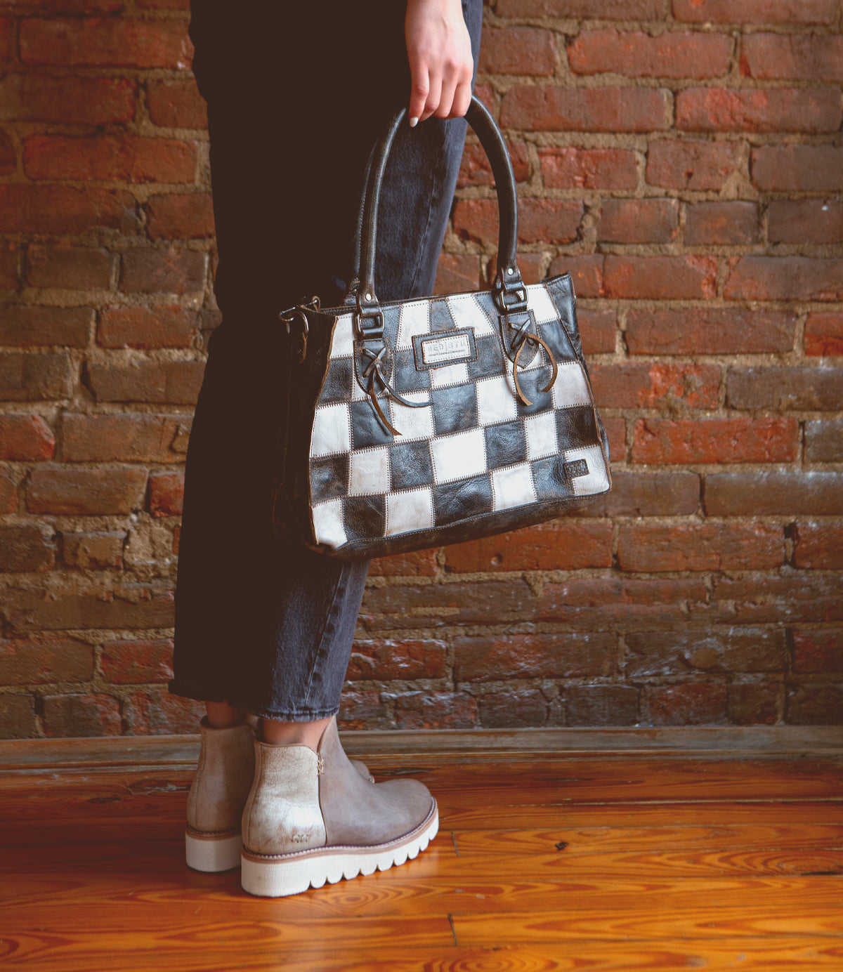 Person holding the Bed|Stü Rockababy SL, a black and white checkered sustainable handbag, wearing dark jeans and taupe ankle boots, standing on a wooden floor in front of a brick wall.