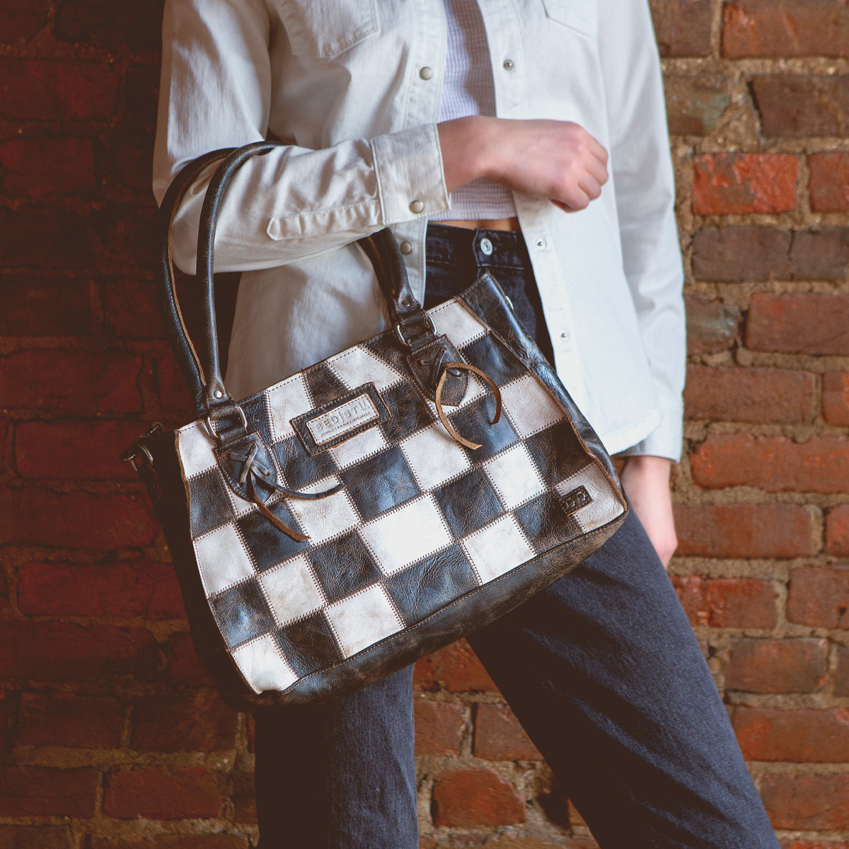 A person in a white shirt and dark jeans holds the Bed|Stü Rockababy SL, a compact crossbody bag crafted from upcycled leather, while standing against a brick wall.