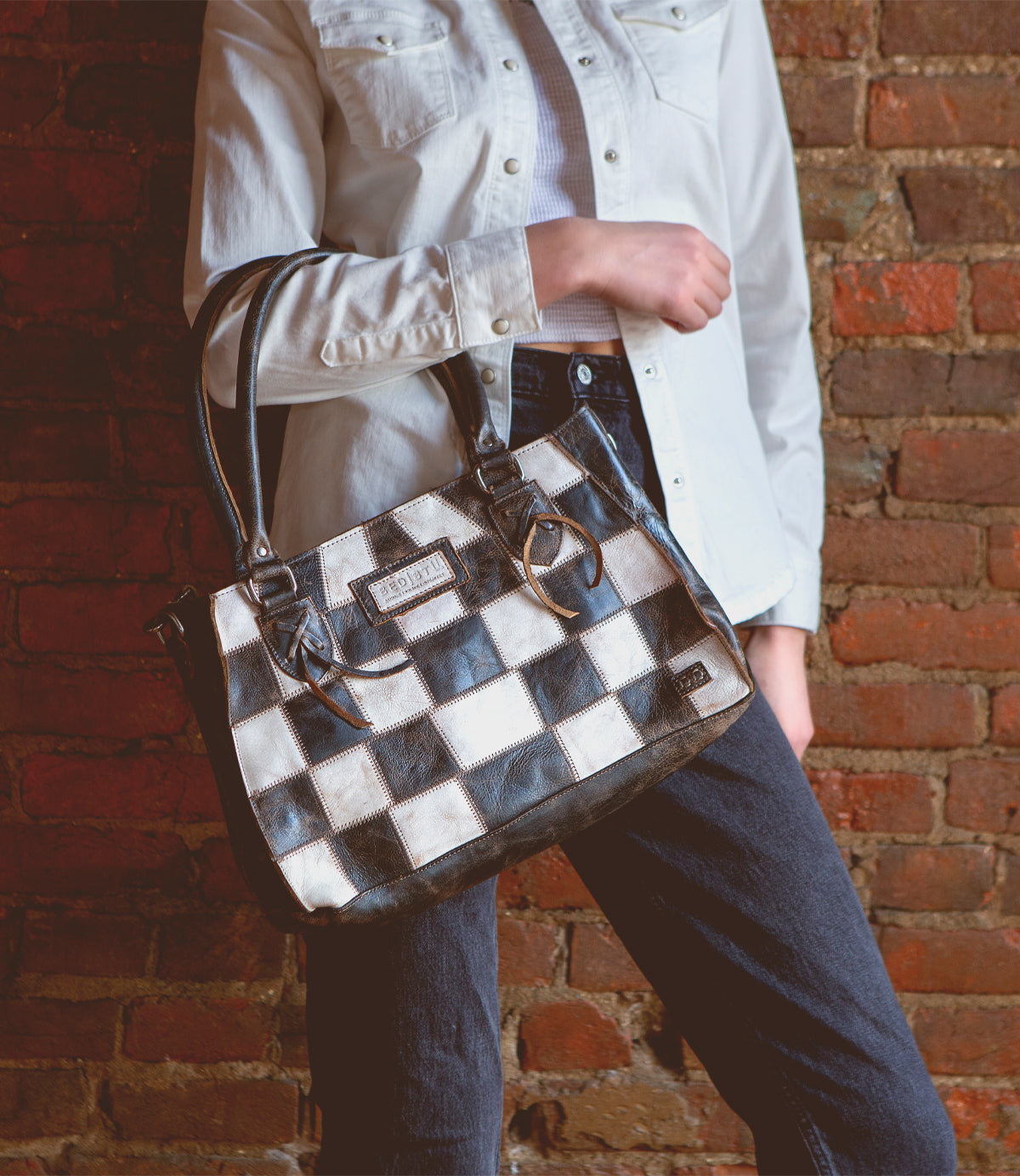 A person in a white shirt and dark jeans holds the Bed|Stü Rockababy SL, a compact crossbody bag crafted from upcycled leather, while standing against a brick wall.
