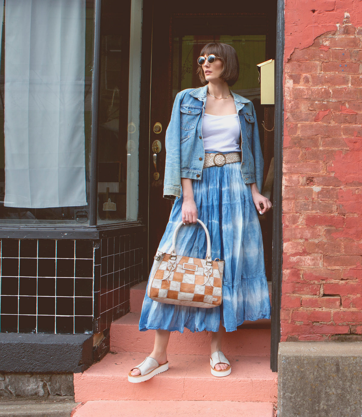 A woman in sunglasses and a denim jacket stands on pink steps holding the Bed|Stü Rockababy SL sustainable handbag, wearing a white top, blue tie-dye skirt, and white sandals in front of a brick building.