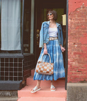 A woman in sunglasses and a denim jacket stands on pink steps holding the Bed|Stü Rockababy SL sustainable handbag, wearing a white top, blue tie-dye skirt, and white sandals in front of a brick building.