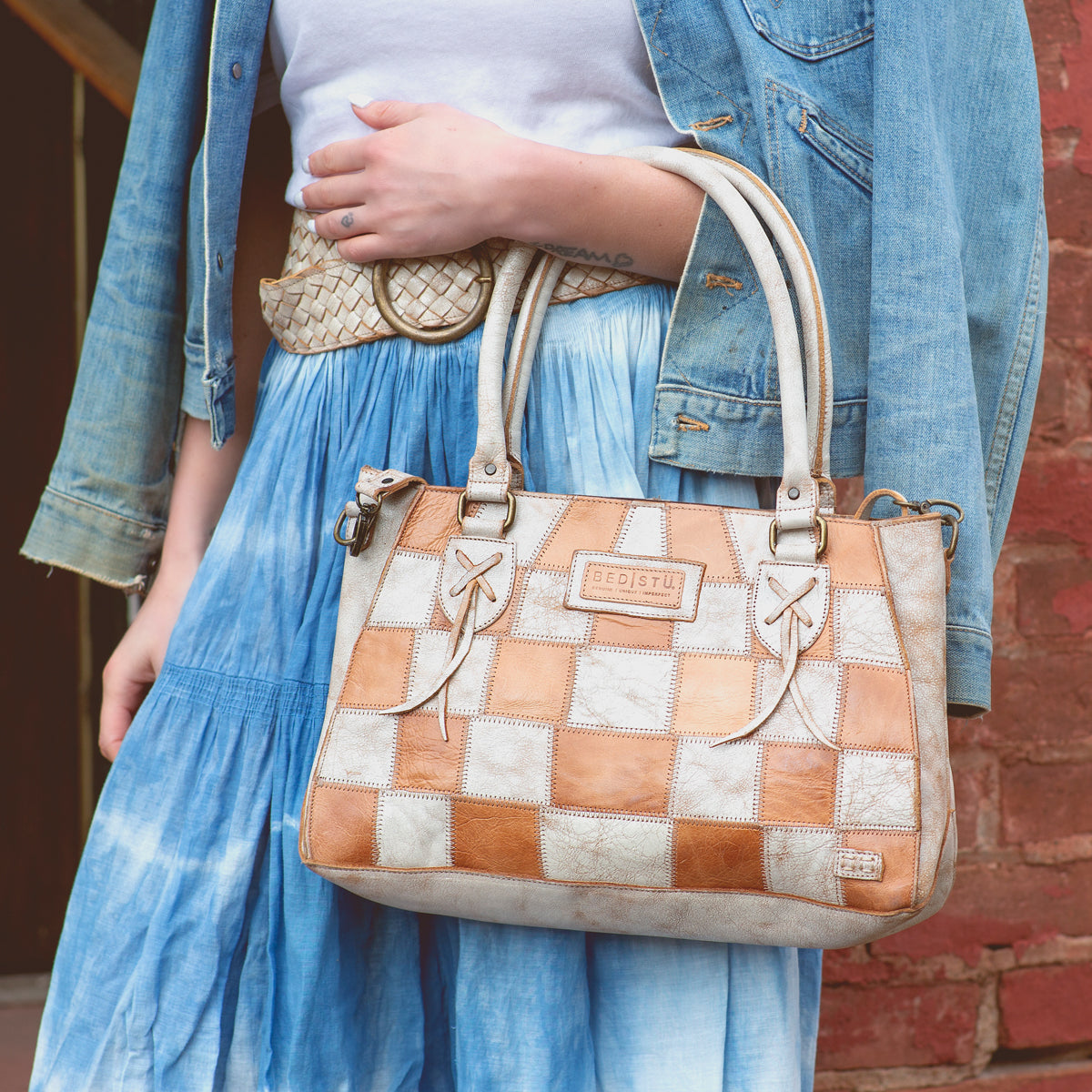 A person wearing a denim jacket and tie-dye skirt holds the Bed|Stü Rockababy SL sustainable handbag, featuring handles and bow details, while standing by a red brick wall.