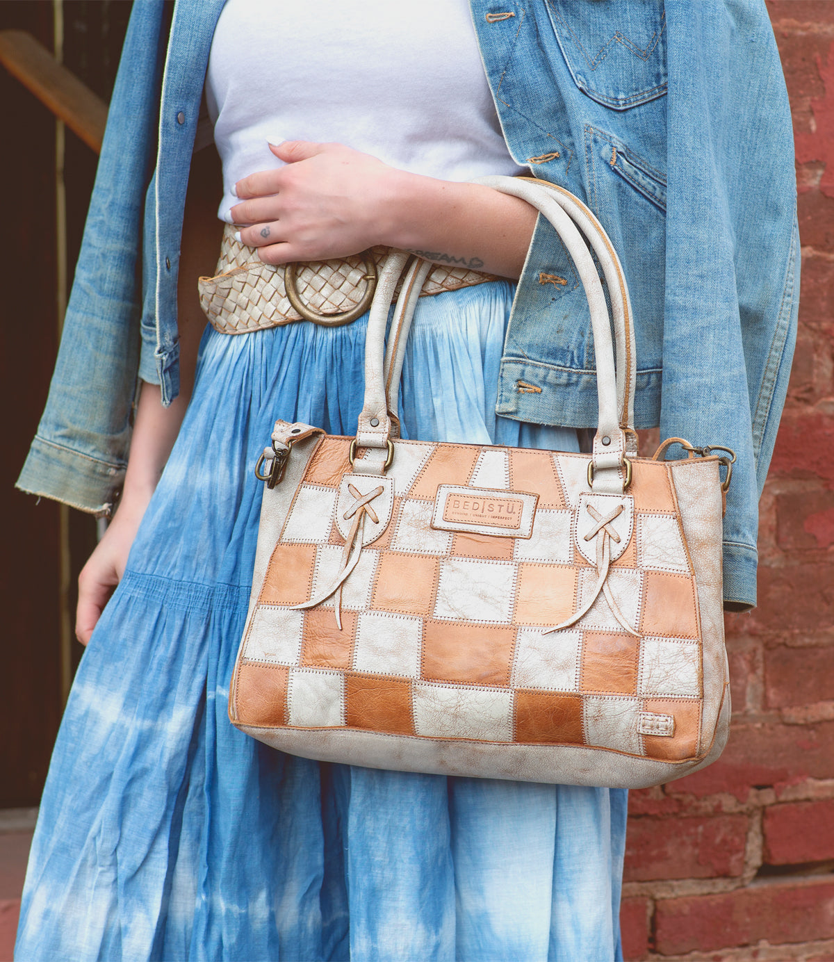 A person wearing a denim jacket and tie-dye skirt holds the Bed|Stü Rockababy SL sustainable handbag, featuring handles and bow details, while standing by a red brick wall.