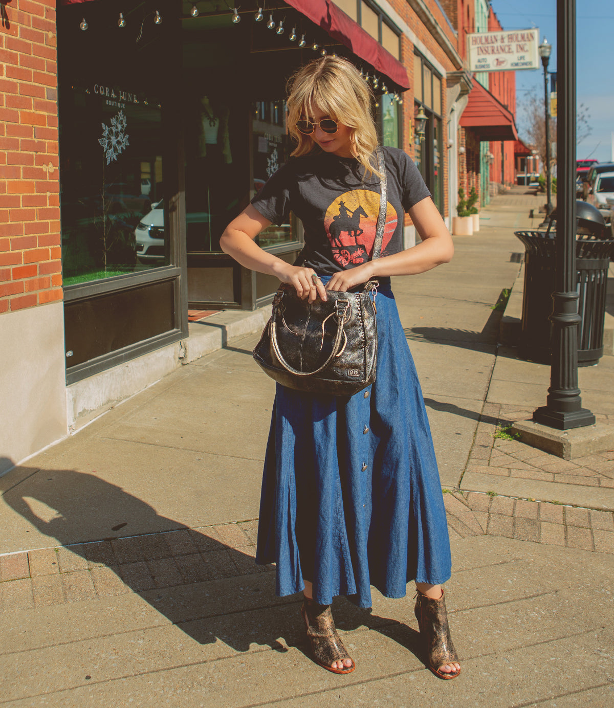 A woman stands on a sunny sidewalk in front of a brick building, looking into her Bed|Stü Rockababy compact leather bag with a crossbody strap, while wearing sunglasses, a printed t-shirt, and a long blue skirt.