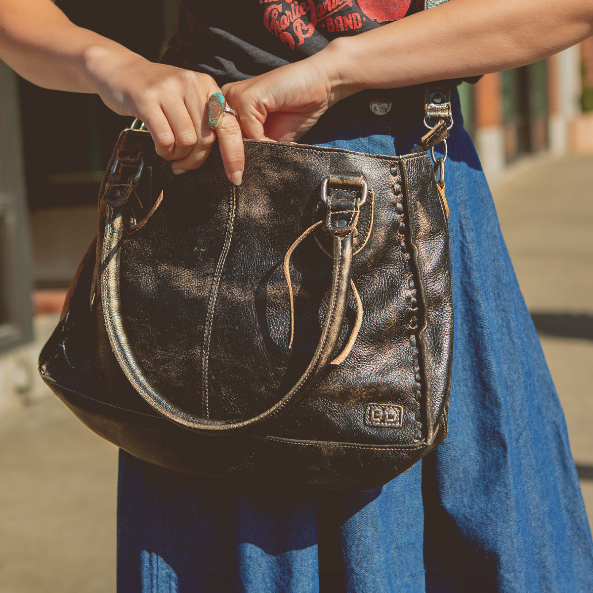 A person wearing a blue skirt and graphic T-shirt holds the Bed|Stü Rockababy, a large black leather handbag featuring silver buckles and a stylish crossbody strap, while standing outdoors.