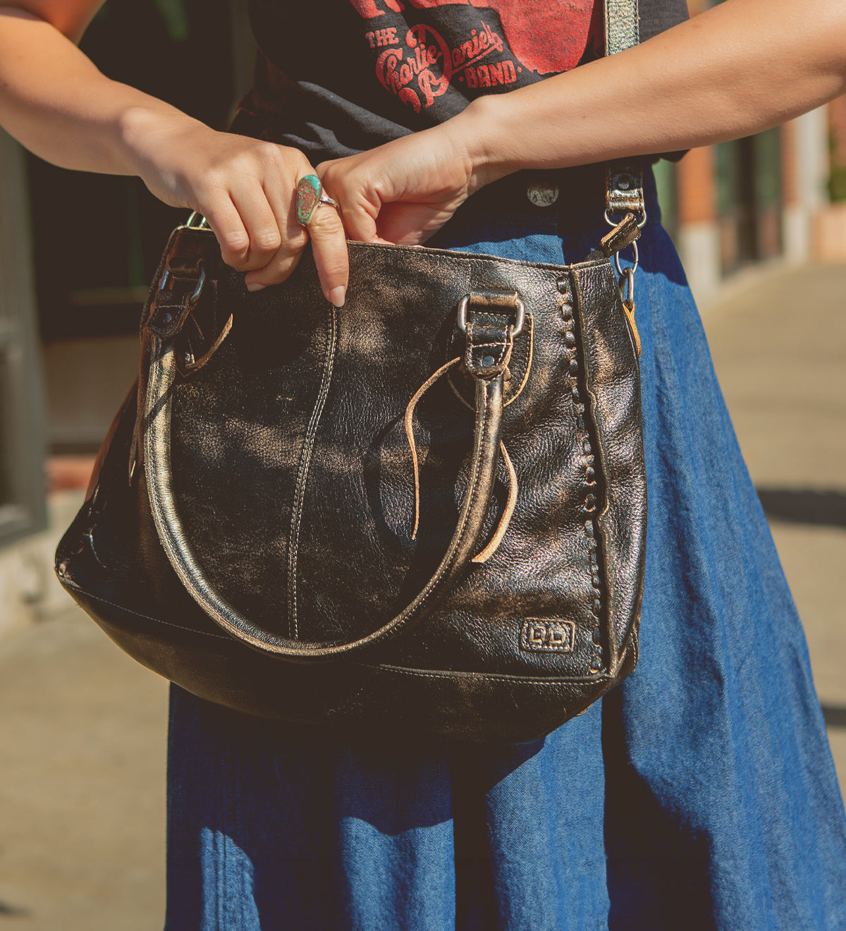 A person wearing a blue skirt and graphic T-shirt holds the Bed|Stü Rockababy, a large black leather handbag featuring silver buckles and a stylish crossbody strap, while standing outdoors.