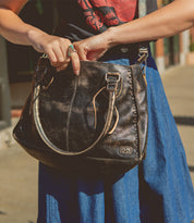 A person wearing a blue skirt and graphic T-shirt holds the Bed|Stü Rockababy, a large black leather handbag featuring silver buckles and a stylish crossbody strap, while standing outdoors.