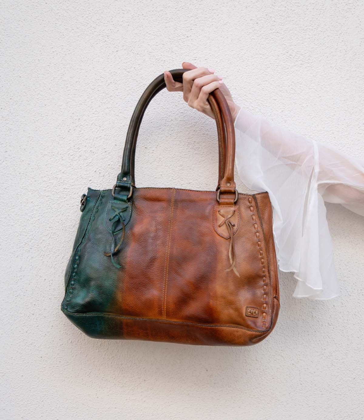 A hand in a sheer white sleeve holds the Bed|Stü Rockababy, a large handbag crafted from vegetable-tanned leather with brown and dark green panels, against a white textured wall.