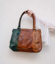 A hand in a sheer white sleeve holds the Bed|Stü Rockababy, a large handbag crafted from vegetable-tanned leather with brown and dark green panels, against a white textured wall.