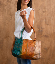 A woman in a sleeveless white top holds the Bed|Stü Rockababy, a large two-tone handbag crafted from vegetable-tanned leather in teal and brown, against a wooden background.