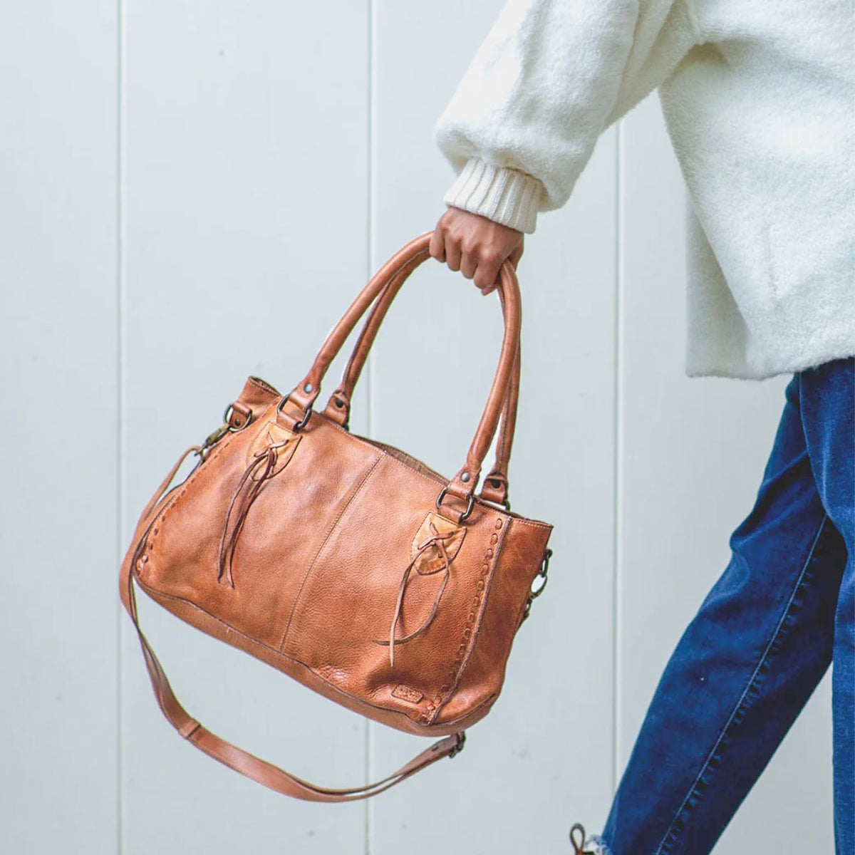 Wearing a white sweater and blue jeans, the person holds the Bed|Stü Rockababy—a compact bag crafted from vegetable-tanned leather with top handles and a crossbody strap—while standing against a light-colored wall.
